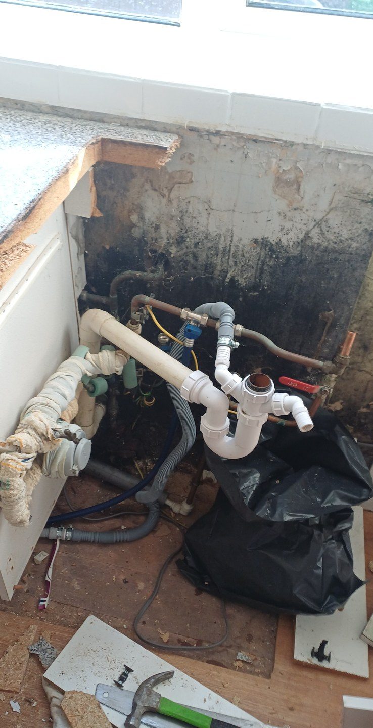 The same kind of room on strip-out day — units out, wall black, floorboards up, pipework exposed.
